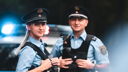Eine junge Polizistin mit ihrem Kollegen vor dem Polizeifahrzeug. Sie blicken in die Kamera