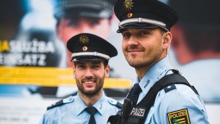 Zwei Polizisten in Uniform lächeln in die Kamera, hinter ihnen das Plakatmotiv der Polizei Sachsen mit einer jungen Kollegin.