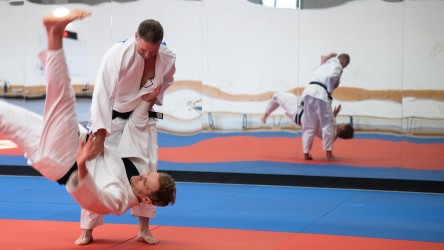 Polizist und Sportler Tobias demonstriert eine Wurftechnik in der Judo-Halle. Ein zweiter Mann, ebenfalls im weißen Judo-Anzug, ist kurz davor, auf der Judo-Matte zu landen.