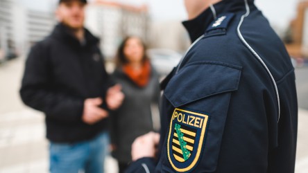 Ein Polizist der Polizei Sachsen im Gespräch mit einem Passanten und einer Passantin. Es sind Schulter und Arm des Polizisten zu sehen. Am Arm hat er das Logo beziehungsweise Wappen der Polizei Sachsen.