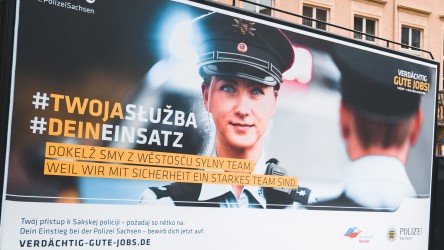 Das Plakatmotiv der Kampagne „Verdächtig gute Jobs“ zeigt eine junge Polizistin in Uniform, welche in die Kamera lächelt. Die Texte auf dem Plakat sind sowohl in sorbischer als auch in deutscher Sprache.